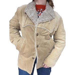 May include: A tan corduroy jacket featuring a cream-coloured, fuzzy collar and lining. The jacket has a button-up front, two pockets, and decorative stitching. The jacket is worn over a red jumper with a geometric pattern.