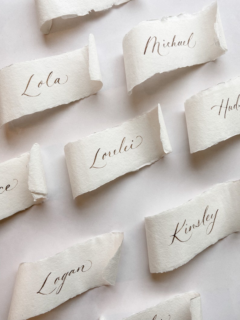 May include: A set of place cards with names written in calligraphy on white paper with a torn edge. The names are Lola, Michael, Hud, Lovelei, Kinsley, and Logan.