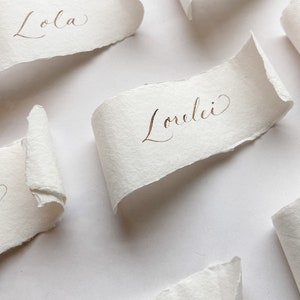 May include: White paper place cards with handwritten names in gold ink. The names are "Lola" and "Lorelei".