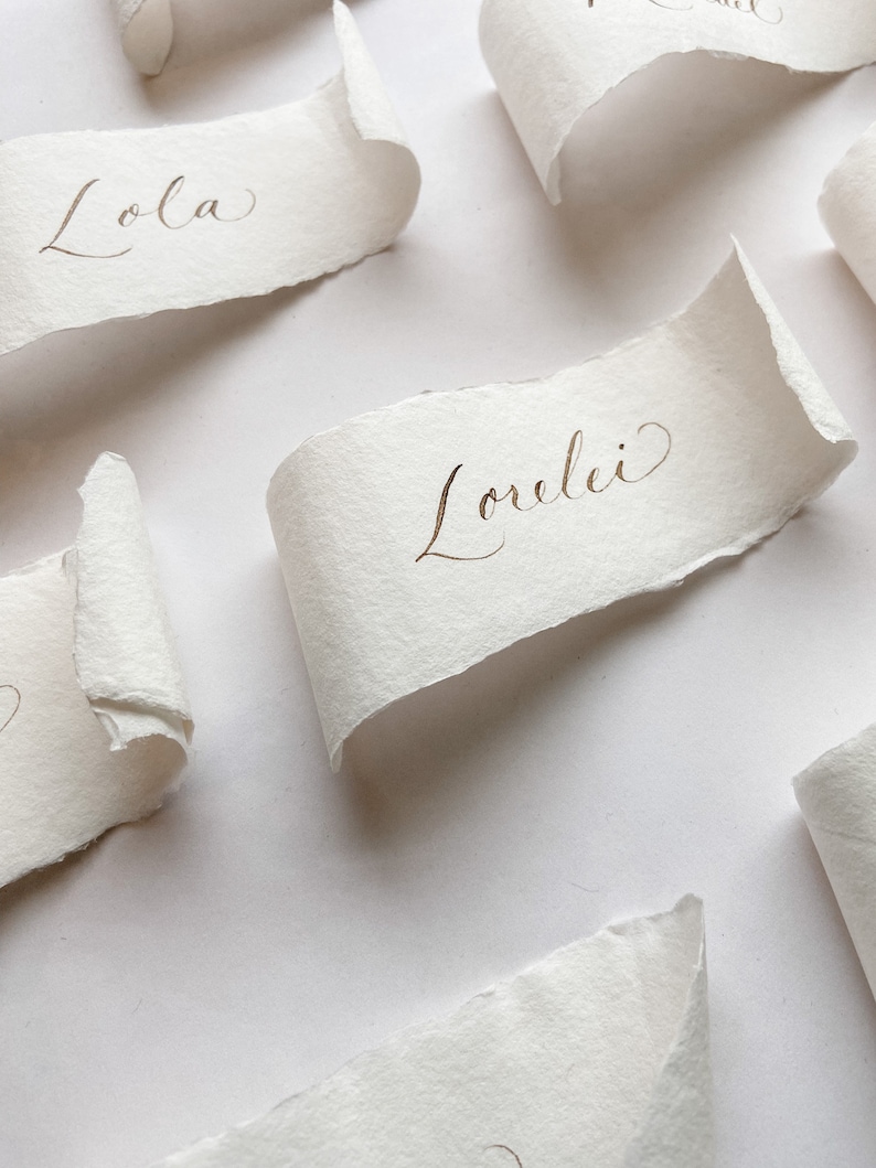 May include: White paper place cards with handwritten names in gold ink. The names are "Lola" and "Lorelei".
