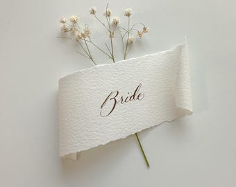May include: A rectangular piece of off-white textured paper with deckled edges, curled at the sides, featuring the word Bride written in brown cursive script. A small sprig of dried white baby's breath flowers is tucked behind the paper.