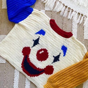 May include: A cream-colored, crocheted sweater with a clown face design. The sweater features blue and gold sleeves, a red collar, and a red, blue, and black clown face. The sweater is handmade and has a textured appearance.