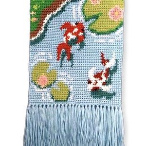 Può includere: Un arazzo da parete in crochet blu con un bordo a frangia. L'arazzo presenta una scena di stagno con ninfee verdi, fiori rosa e pesci rossi e bianchi.