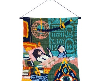 May include: A colorful crocheted wall hanging with a green, blue, and orange color scheme. The hanging features a variety of geometric shapes and patterns, including a large eye in the center.