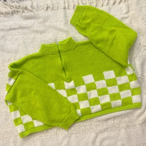 May include: A neon green sweater with a half-zip collar and long sleeves. The bottom of the sweater features a checkerboard pattern in white and green. The sweater is made of a knitted material.