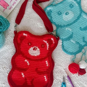 May include: A red, crocheted gummy bear-shaped crossbody bag with a matching strap. The bag features white highlights and a white face. A blue crocheted gummy bear, yarn, scissors, and a crochet hook are also visible.