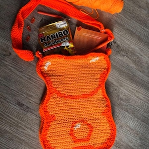 May include: An orange crochet bag in the shape of a gummy bear, with a matching strap. Inside the bag are a packet of Haribo Goldbears sweets, a crochet hook, and a small book. A ball of orange yarn is also visible.