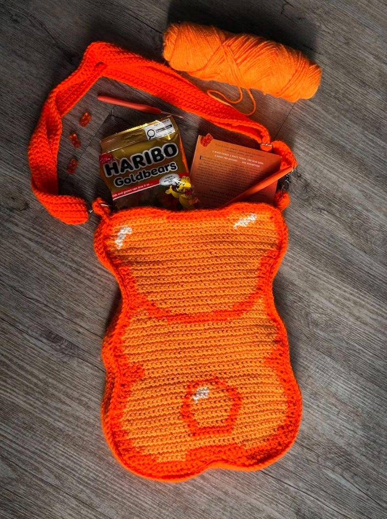 May include: An orange crochet bag in the shape of a gummy bear, with a matching strap. Inside the bag are a packet of Haribo Goldbears sweets, a crochet hook, and a small book. A ball of orange yarn is also visible.
