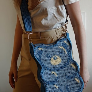 May include: A blue crochet teddy bear-shaped crossbody bag with a matching strap. The bag has a light yellow face and white accents. The bag is worn with a white t-shirt with a brown teddy bear design.