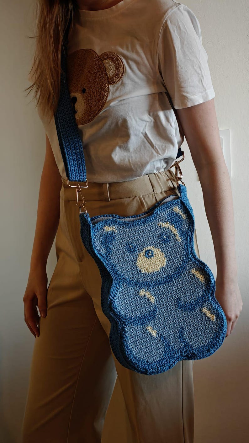 May include: A blue crochet teddy bear-shaped crossbody bag with a matching strap. The bag has a light yellow face and white accents. The bag is worn with a white t-shirt with a brown teddy bear design.