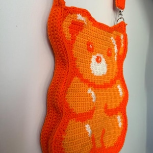 May include: An orange crochet crossbody bag shaped like a gummy bear. The bag features a white and orange design, with a matching orange strap and silver hardware. The bag is made of a soft, textured material.
