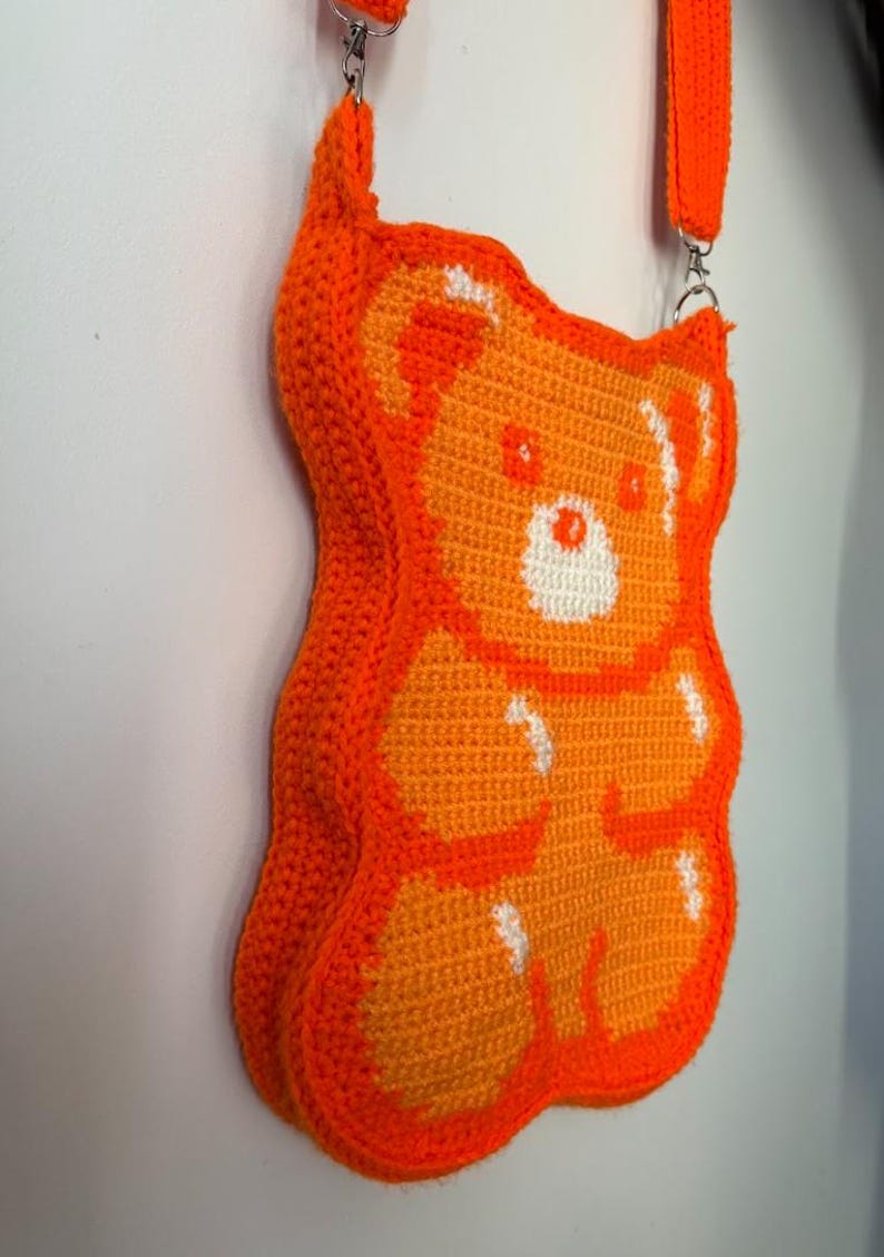 May include: An orange crochet crossbody bag shaped like a gummy bear. The bag features a white and orange design, with a matching orange strap and silver hardware. The bag is made of a soft, textured material.