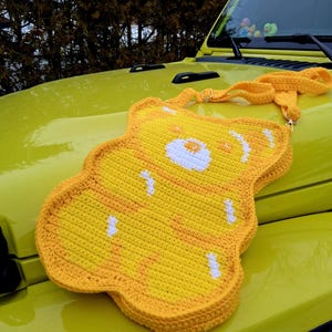 May include: A yellow crochet crossbody bag in the shape of a bear, featuring white and orange details. The bag, with its long strap, is shown on the bonnet of a yellow vehicle. The bear's face is detailed with white and orange stitching.