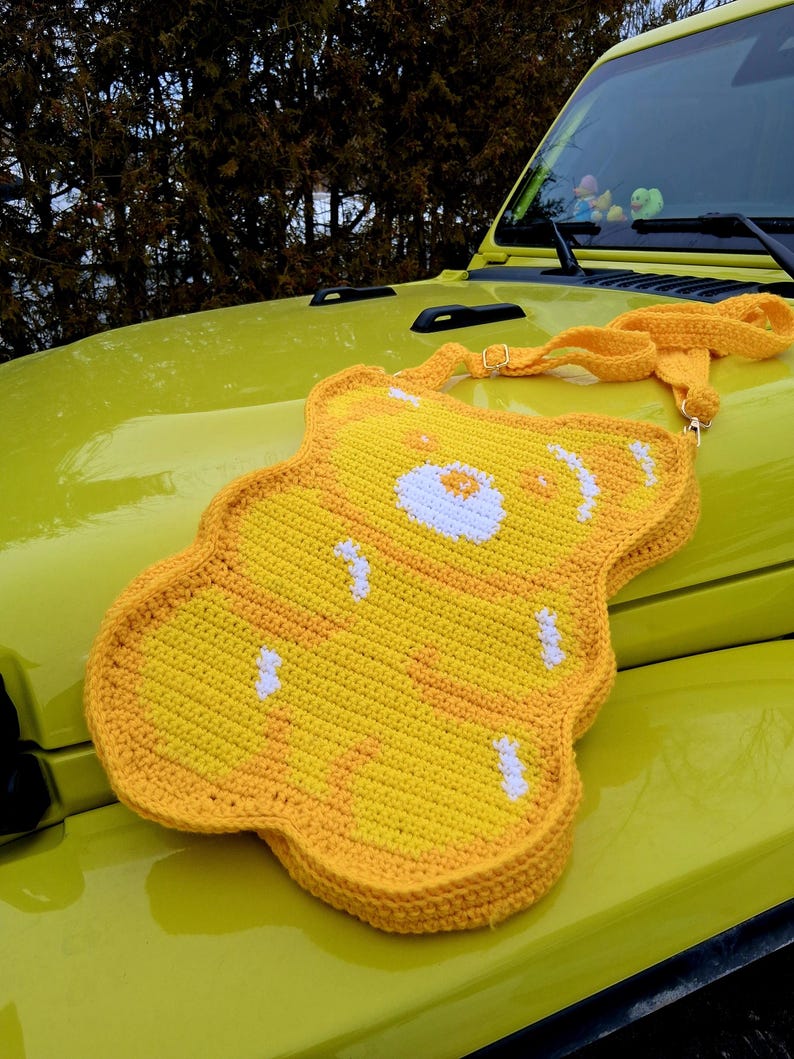 May include: A yellow crochet crossbody bag in the shape of a bear, featuring white and orange details. The bag, with its long strap, is shown on the bonnet of a yellow vehicle. The bear's face is detailed with white and orange stitching.