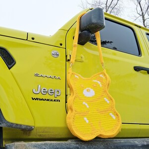 May include: A yellow crochet bag shaped like a gummy bear, with white detailing. The bag is suspended by a yellow strap, set against a yellow Jeep Wrangler. The design of the bag is a fun and quirky accessory.