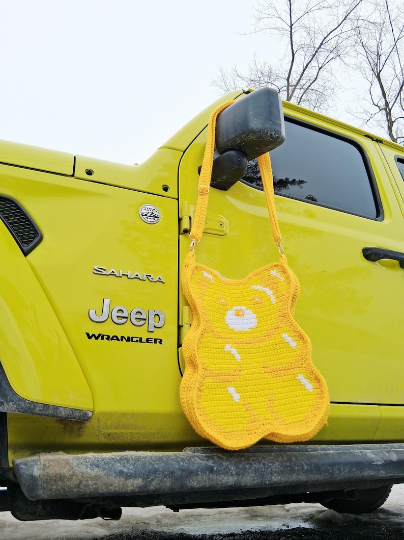 May include: A yellow crochet bag shaped like a gummy bear, with white detailing. The bag is suspended by a yellow strap, set against a yellow Jeep Wrangler. The design of the bag is a fun and quirky accessory.