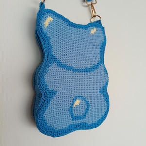 May include: A handmade, light blue crochet crossbody bag shaped like a gummy bear. The bag has a darker blue outline and strap, with gold-coloured metal hardware. Small yellow accents are visible on the bag's surface.