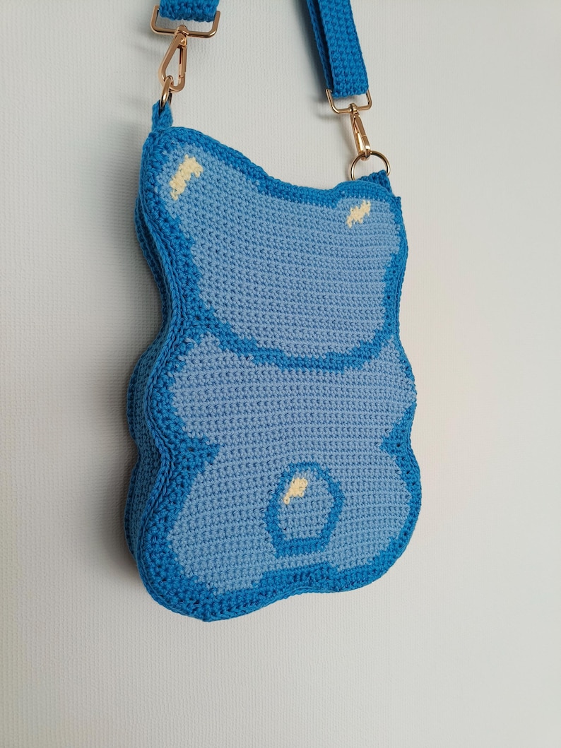 May include: A handmade, light blue crochet crossbody bag shaped like a gummy bear. The bag has a darker blue outline and strap, with gold-coloured metal hardware. Small yellow accents are visible on the bag's surface.