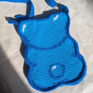 May include: A handmade, blue crochet crossbody bag with a unique, rounded shape. The bag features a darker blue border and strap, with small white accents. The strap is attached with silver-coloured metal rings. The bag is designed with a textured, woven pattern.