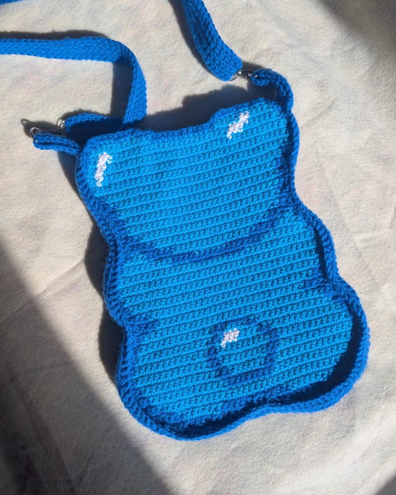 May include: A handmade, blue crochet crossbody bag with a unique, rounded shape. The bag features a darker blue border and strap, with small white accents. The strap is attached with silver-coloured metal rings. The bag is designed with a textured, woven pattern.