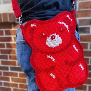 May include: A red, crocheted crossbody bag in the shape of a gummy bear. The bag features a white face and white accents, with a red strap and silver hardware. The bag is worn over the shoulder.