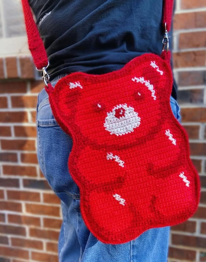 May include: A red, crocheted crossbody bag in the shape of a gummy bear. The bag features a white face and white accents, with a red strap and silver hardware. The bag is worn over the shoulder.