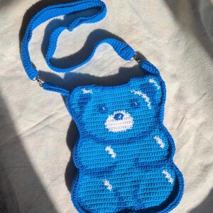 May include: A handmade, blue crochet crossbody bag shaped like a gummy bear. The bag has white detailing and a matching blue strap. The bag is made from soft, textured yarn and has a playful, whimsical design.