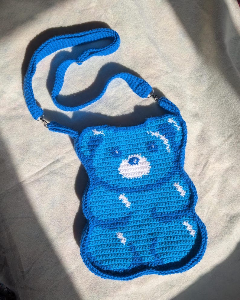 May include: A handmade, blue crochet crossbody bag shaped like a gummy bear. The bag has white detailing and a matching blue strap. The bag is made from soft, textured yarn and has a playful, whimsical design.