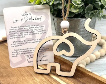 May include: A wooden elephant ornament with a cutout heart shape. The ornament is hanging from a twine string with a white bead. A card with the text "A Story of Love & Sisterhood" is in the background.