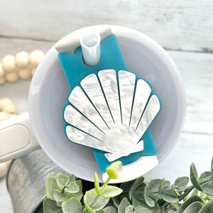 May include: A teal and white seashell-shaped drink accessory sits atop a white tumbler. The seashell design has a pearlescent finish. The tumbler has a white straw and a white lid. The background includes greenery and a metal bucket.