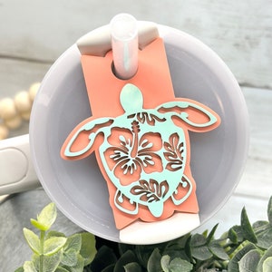 May include: A light pink and blue plastic turtle with a hibiscus flower design. The turtle is attached to a straw lid for a reusable cup.