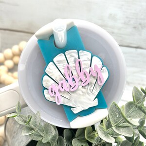 May include: A turquoise blue lid with a white seashell design and the word "gabby" written in pink. The lid is attached to a white tumbler.