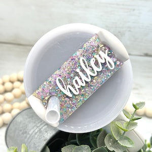 May include: A white cup with a clear straw and a glittery pink, purple, and green name tag that says "Hailey".