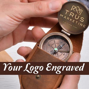 May include: A vintage-style compass with a bronze finish, held in a brown leather case. The compass features a detailed dial and is open, revealing the needle. The text "Your Logo Engraved" is displayed.