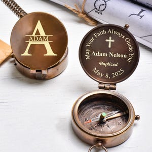 May include: Two antique brass compasses. One is closed, engraved with the letter "A" and the name "ADAM". The other is open, revealing the compass dial, and has the inscription "May Your Faith Always Guide You" with a cross, name, and date.
