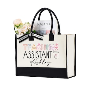 May include: White canvas tote bag with black trim and handles. The bag has the text "TEACHING ASSISTANT Ashley" printed on it in black and colorful fonts. The text is surrounded by colorful school supplies.