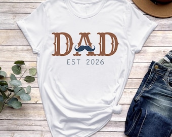 May include: White t-shirt with the word "DAD" in brown, stylized lettering and a blue mustache graphic. Below the word "DAD" is the text "EST 2026". The shirt is folded on a wooden surface.