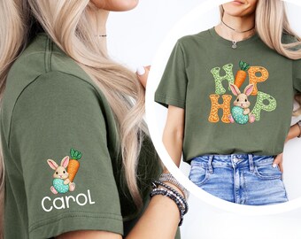 Peut inclure: T-shirt vert olive avec un motif coloré "Hip Hop" représentant un lapin tenant un œuf de Pâques et des carottes. La manche gauche porte un motif de lapin et de carotte avec le nom "Carol". Un haut décontracté et festif.
