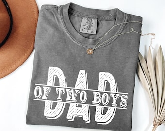 May include: A gray t-shirt with the text "DAD OF TWO BOYS" in white, distressed lettering. A gold necklace with a small pendant rests on the shirt. The shirt is folded, and a brown hat is partially visible in the background.
