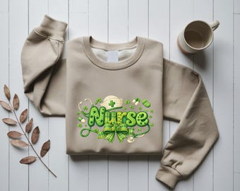 Peut inclure: Un sweat-shirt beige avec un motif vert et blanc "Infirmière". Le motif comprend un chapeau d'infirmière, un stéthoscope, des trèfles et un nœud. Une tasse blanche est en arrière-plan.