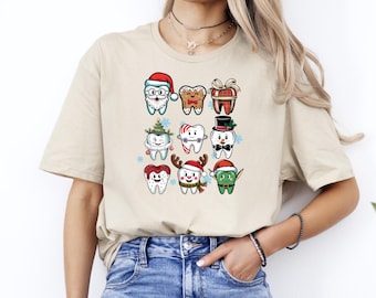 May include: Beige t-shirt featuring a festive graphic of cartoon teeth dressed in Christmas attire. The teeth are decorated as Santa, gingerbread, presents, and more. The shirt is a comfortable, casual style.