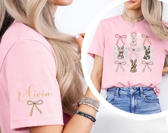 Peut inclure: T-shirt rose clair avec un motif de lapins et de nœuds. Les lapins ont des motifs floraux et à motifs. La manche porte le nom "Olivia" en lettres dorées avec un motif de nœud. Le modèle porte un jean bleu.
