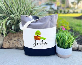 May include: A grey and black tote bag with a white panel featuring a green worm reading a book. The worm is wearing glasses and the text "Jenifer" is printed below the worm. The bag is a tri-color grey.