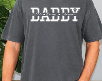 May include: Dark gray t-shirt with the word "DADDY" in a white, collegiate-style font. Below the word "DADDY" are the names Adele, Emma, and Angelina. The shirt is a solid color and has short sleeves.