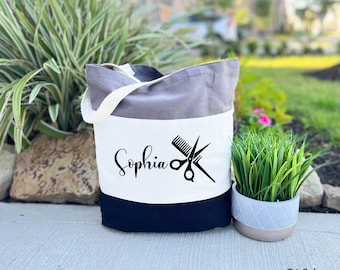 May include: A tri-color tote bag with a gray, white, and black design. The bag has a black and white graphic of scissors and a comb with the name "Sophia" printed on it.