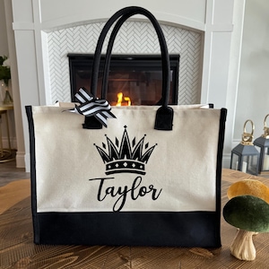 May include: Cream-colored tote bag with black accents, featuring a black crown design and the name "Taylor". The bag has black handles and a black and white striped bow. The bottom of the bag is black.