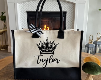 May include: Cream-colored tote bag with black accents, featuring a black crown design and the name "Taylor". The bag has black handles and a black and white striped bow. The bottom of the bag is black.