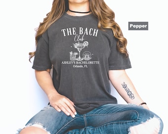 May include: A dark gray t-shirt with the text "THE BACH Club" and "ASHLEY'S BACHELORETTE Orlando, FL" in white. The shirt features a cocktail and seashell graphic. The shirt is a Pepper color.