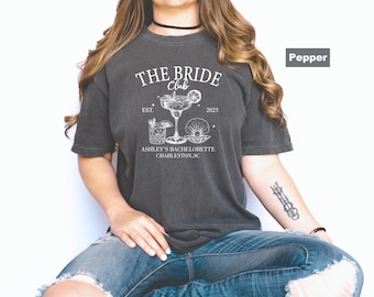 May include: A dark gray t-shirt with the text "THE BRIDE CLUB" and the year "2025". The shirt features a graphic of a cocktail, a drink, and a seashell. The text "ASHLEY'S BACHELORETTE CHARLESTON, SC" is also printed on the shirt.