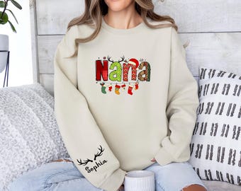 Peut inclure: Sweat-shirt crème avec le mot "Nana" dans un design festif et coloré. Les lettres sont décorées de motifs de Noël et d'un bonnet de Père Noël. La manche présente un motif de bois de cerf et le nom "Sophia".